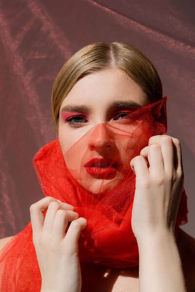 Fair haired model with red eye shadow touching tulle cloth on grey background 