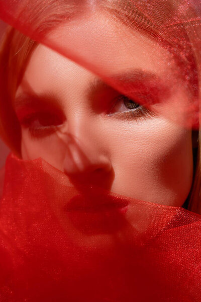 Close up view of young woman looking at camera near red blurred cloth 