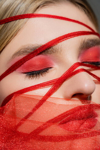 Close up view of woman with red eye shadow, threads and fabric near face isolated on grey 