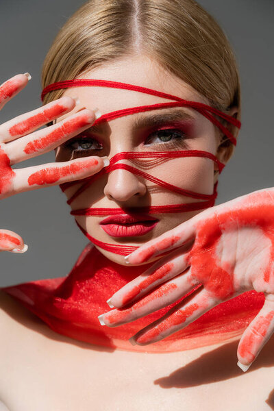 Young model with red threads on face and paint on hands looking at camera isolated on grey 