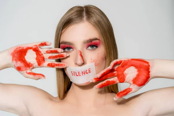 Young woman with paint on hands and lettering silence on paper on mouth isolated on grey