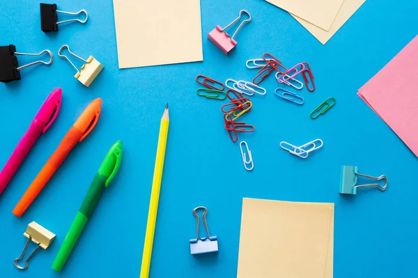 top view of empty paper notes near colorful pens and paper clips on blue