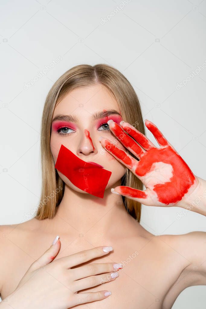 Young woman with red adhesive tape on mouth and paint on hand isolated on grey 