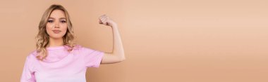 smiling woman looking at camera and showing muscles isolated on beige, banner