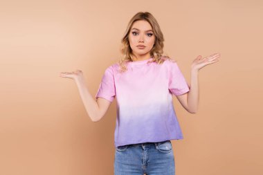 confused woman showing shrug gesture and looking at camera isolated on beige clipart