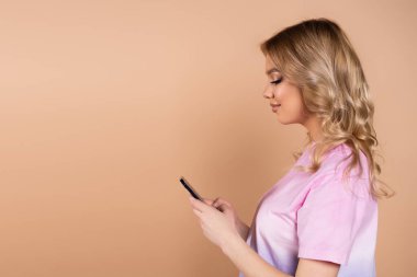 Dalgalı saçlı, akıllı telefondan mesaj atan gülümseyen kadının yan görünüşü bejde izole edilmiş.