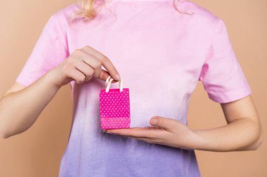 cropped view of woman in t-shirt holding tiny gift bag isolated on beige