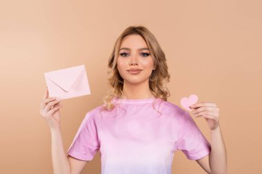 happy woman with wavy hair holding envelope and paper heart isolated on beige