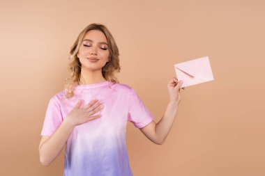 pleased woman with closed eyes holding postal envelope isolated on beige