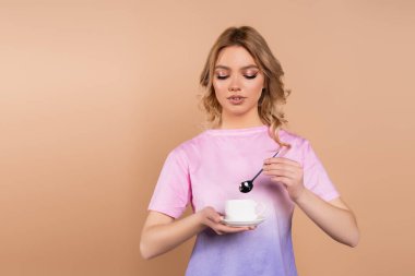 Dalgalı saçlı, elinde kahve fincanı ve bej rengi çay kaşığı tutan genç bir kadın.
