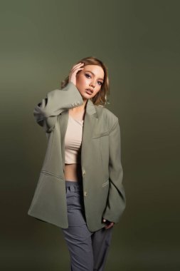 stylish woman in grey oversize blazer touching wavy hair isolated on green