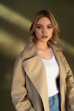 woman with wavy hair posing in trench coat and looking at camera on beige background