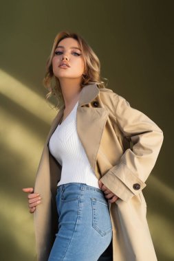 Trençkotlu ve kot pantolonlu güzel bir kadının bej arka planda kameraya bakışının düşük açılı görüntüsü.