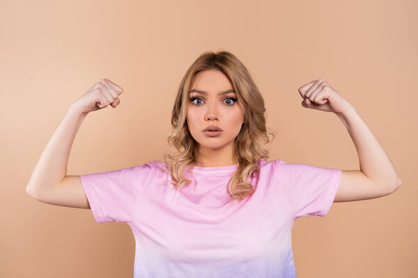 young woman showing muscles and looking at camera isolated on beige