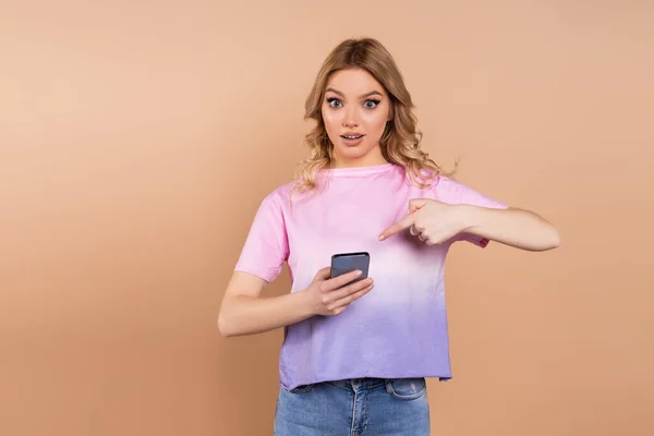 Überraschte Frau Zeigt Auf Handy Und Blickt Vereinzelt Auf Beige — Stockfoto