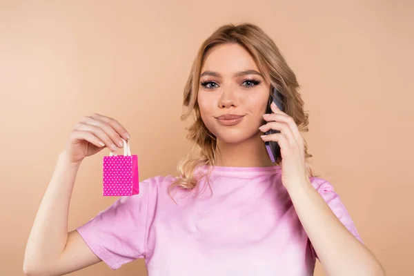 cheerful woman holding tiny polka dot shopping bag while talking on cellphone isolated on beige