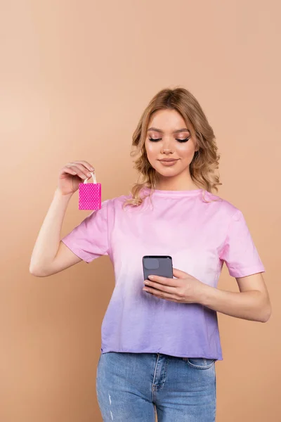 pleased woman holding smartphone and tiny shopping bag isolated on beige