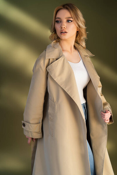 woman with wavy hair looking away while posing in trench coat on beige background