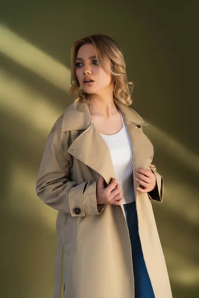 sensual young woman in trench coat looking away on beige background