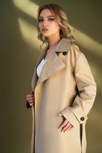 stylish woman in trench coat looking at camera on beige background