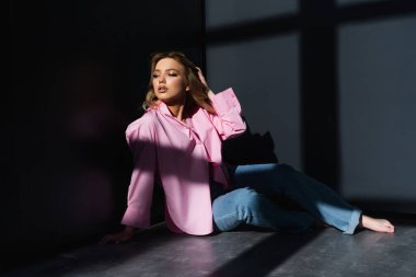 Pembe gömlekli çıplak ayaklı bir kadın siyah duvarların yanında gölgeli bir şekilde oturuyor.