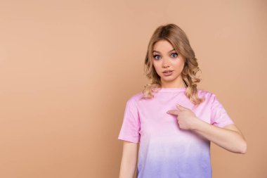 Şaşırmış bir kadın kendini işaret ederek bej rengi bir elbiseyi işaret ediyor.