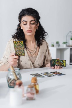 KYIV, UKRAINE - FEBRUARY 23, 2022: Gypsy fortune teller holding tarot cards near blurred witchcraft supplies at home 