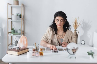 Gypsy fortune teller holding tarot cards near blurred orb on table 