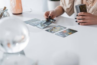 KYIV, UKRAINE - FEBRUARY 23, 2022: Cropped view of fortune teller holding tarot cards near blurred orb on table 