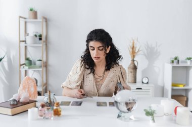 Gypsy fortune teller holding tarot cards near candles and witchcraft supplies on table 