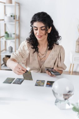 Positive gypsy fortune teller holding blurred tarot cards at home 
