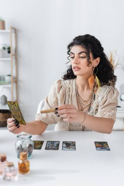 KYIV, UKRAINE - FEBRUARY 23, 2022: Young gypsy fortune teller holding tarot card and sage stick at home 