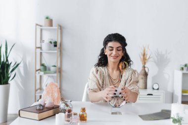 Smiling gypsy fortune teller touching magic orb near crystals at home 