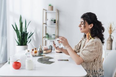 KYIV, UKRAINE - FEBRUARY 23, 2022: Side view of positive gypsy soothsayer holding orb near witchcraft supplies on table at home 