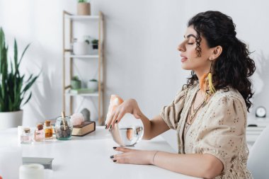 Side view of gypsy soothsayer holding magic orb near jars on table 