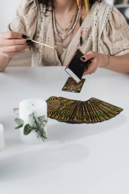 KYIV, UKRAINE - FEBRUARY 23, 2022: Cropped view of fortune teller holding match near tarot cards and candle on table 