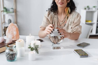 KYIV, UKRAINE - FEBRUARY 23, 2022: Cropped view of smiling fortune teller touching orb near tarot and candles 