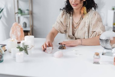 Cropped view of smiling fortune teller holding magic crystals near candles and orb at home 