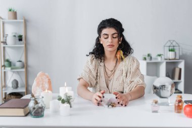 Gypsy soothsayer touching magic crystals near book, orb and candles on table 