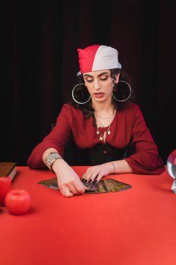 Gypsy soothsayer holding tarot cards near orb and book on table isolated on black 