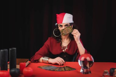 KYIV, UKRAINE - FEBRUARY 23, 2022: Gypsy fortune teller holding tarot card near face and witchcraft supplies on table isolated on black 