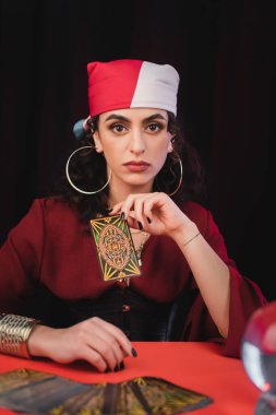 KYIV, UKRAINE - FEBRUARY 23, 2022: Gypsy fortune teller holding tarot card near blurred magic orb on table isolated on black 