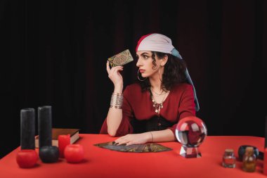 KYIV, UKRAINE - FEBRUARY 23, 2022: Young gypsy fortune teller holding tarot near witchcraft supplies on table isolated on black 