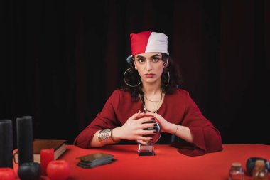 Gypsy fortune teller touching magic orb near tarot and candles isolated on black 
