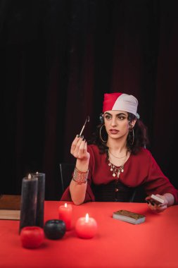 Gypsy medium holding match near tarot cards and candles isolated on black 
