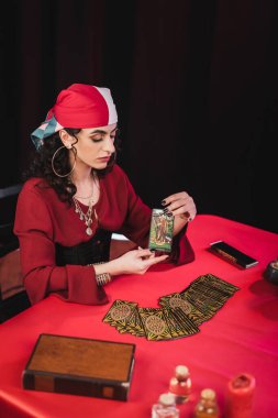 KYIV, UKRAINE - FEBRUARY 23, 2022: Young gypsy fortune teller holding tarot card near jars and book isolated on black  clipart