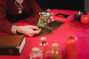 KYIV, UKRAINE - 23 Şubat 2022: Kâhin 'in kitap yanında tarot kartı tutuşu ve siyah masanın üzerindeki bulanık kavanozlar 
