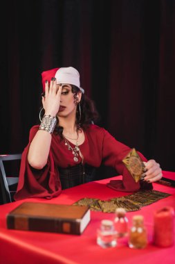 Scared gypsy medium holding tarot card near book on black 