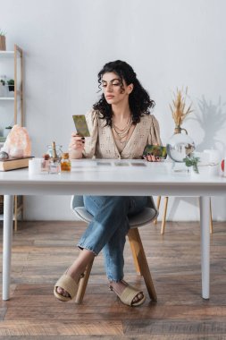 Gypsy soothsayer holding blurred tarot cards near witchcraft supplies on table 