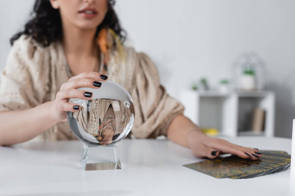 Cropped view of blurred fortune teller touching orb near tarot cards at home 